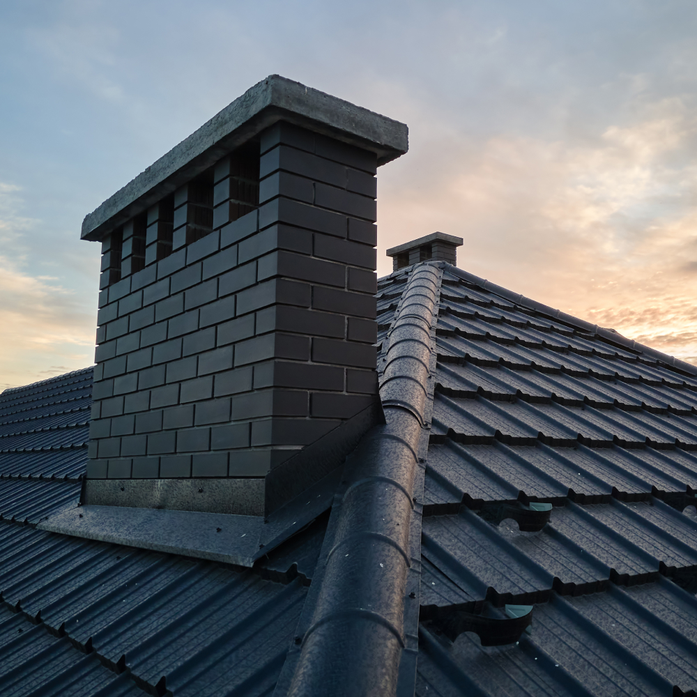 Dach domu pokryty ciemną dachówką ceramiczną z widocznym murowanym kominem o zachodzie słońca.